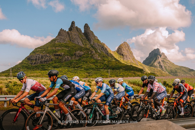 The Tour de Maurice peloton rides in front of a series of three jagged peaks, with vegetation at teh base and rocky ridges at the top.